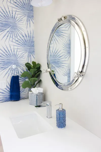 The bathroom boasts a nautical theme with a porthole mirror and blue palm wallpaper. Modern fixtures and crisp design elements create a refreshing and unique space.
